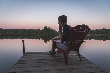 Young man on his cell phone while watching the sunset from a chair on a dock.