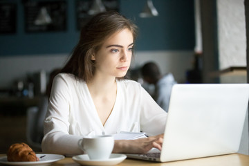 Focused beautiful young girl work at laptop sitting in cafe with coffee and croissant, serious...