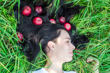 Naklejka premium Charming girl the brunette lies on a green grass on a glade. Hair on the ground. Red apples in hair. Summer mood memories. Natural nutrition, health.