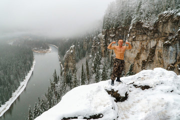 Naklejka na ściany i meble Strong happy young man with bare torso on mountain top covered with snow celebrating success and showing muscles