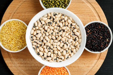 Set of different cereals and legumes in a white bowls on a black table, top view