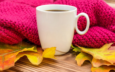 Cup of coffee with autumn leaves and knitted scarf on wooden background.