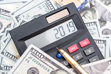 Calculator with numbers 2019 and pen on background of dollar bills.