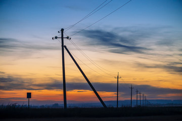 Fototapeta na wymiar Power lines in field on sunrise background. Silhouettes of poles with wires at dawn. Cables of high voltage on warm orange blue sky. Power industry at sunset. Multicolored picturesque vivid sky.