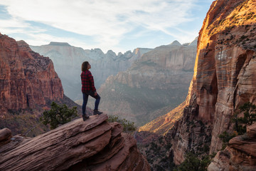 Fototapeta premium Adventurous Girl at the edge of a cliff is looking at a beautiful landscape view in the Canyon during a vibrant sunset. Taken in Zion National Park, Utah, United States.