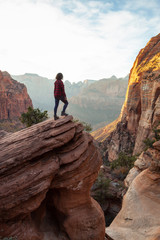 Adventurous Girl at the edge of a cliff is looking at a beautiful landscape view in the Canyon during a vibrant sunset. Taken in Zion National Park, Utah, United States.