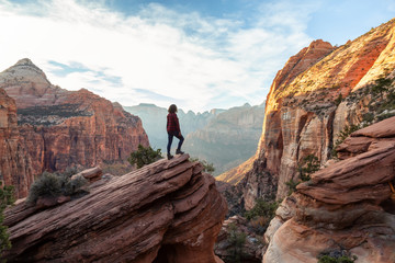 Adventurous Woman at the edge of a cliff is looking at a beautiful landscape view in the Canyon during a vibrant sunset. Taken in Zion National Park, Utah, United States. - obrazy, fototapety, plakaty