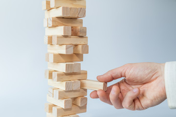 Man's hand taking or putting a block to an unstable and incomplete tower of wooden blocks. Concept photo of planning, taking risks and strategizing. Light gray background.