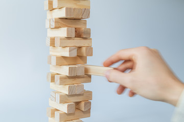 Man's hand taking or putting a block to an unstable and incomplete tower of wooden blocks. Concept photo of planning, taking risks and strategizing. Hand is in motion. Light gray background.