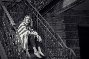Black and white photo, portrait of a young sad woman sitting on a stone fence near the house. Depressed.