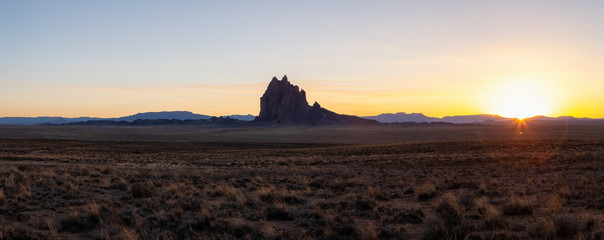 Striking panoramic landscape view of a dry desert with a mountain peak in the background during a vibrant sunset. Taken at Shiprock, New Mexico, United States.