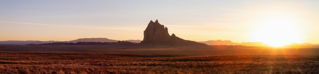 Fototapeta na wymiar Striking panoramic landscape view of a dry desert with a mountain peak in the background during a vibrant sunset. Taken at Shiprock, New Mexico, United States.