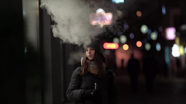 A girl is smoking vape outside in the evening. Nice colorful bokeh in front of the evening city.