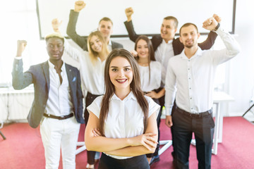 Young white female executive standing with crossed hands in front of colleagues with their arms raised up celebrate win.