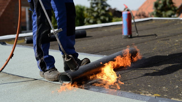Dachdecker auf Flachdach Baustelle in der Industrie macht Dach neu: Isolierter Flachdach Geselle schwei&szlig;t industriell Flachdach Dachpappe in Handarbeit mit offener Flamme  auf flaches Dach