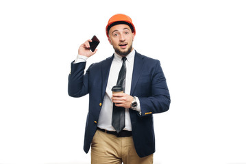 Business portrait of surprised contractor and developer, talking about phone.  Businessman in hard hat with cup of coffee standing over white background. News and coffee brake concept