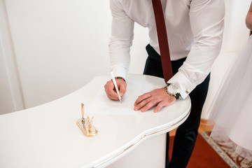 a man in a white shirt with a burgundy red tie signs a paper with a ballpoint pen on a white table closeup