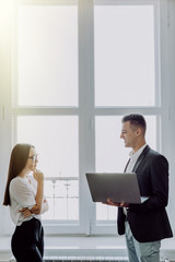 Happy couple of young business partners working in modern office. Two coworkers working on laptop while standing near window, copy space