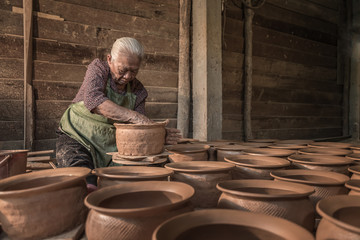 A woman potter in a plaid shirt and green apron beautifully sculpts a deep bowl of brown clay and cuts off excess clay on a potter's wheel