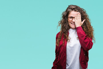 Beautiful brunette curly hair young girl wearing jacket and glasses over isolated background covering one eye with hand with confident smile on face and surprise emotion.