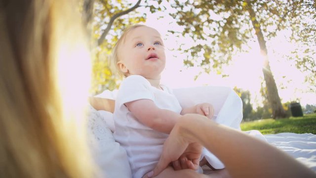 Cute blonde young Caucasian mother lying outdoors on the blanket watching her baby son playing 
