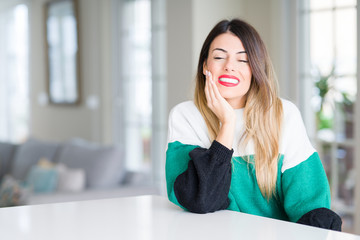 Obraz premium Young beautiful woman wearing winter sweater at home touching mouth with hand with painful expression because of toothache or dental illness on teeth. Dentist concept.