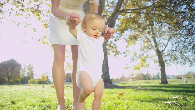 Happy baby girl supported by her young Caucasian mom as she practices walking on the grass 