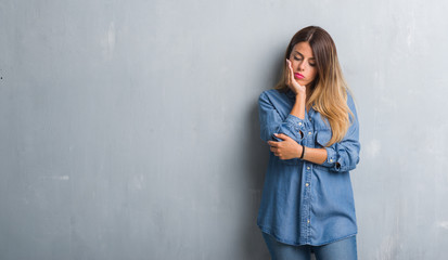 Young adult woman over grunge grey wall wearing denim outfit thinking looking tired and bored with depression problems with crossed arms.