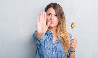 Young adult woman over grey grunge wall eating lollipop candy with open hand doing stop sign with serious and confident expression, defense gesture