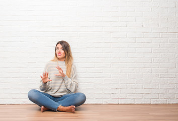 Young adult woman sitting on the floor over white brick wall at home disgusted expression, displeased and fearful doing disgust face because aversion reaction. With hands raised. Annoying concept.