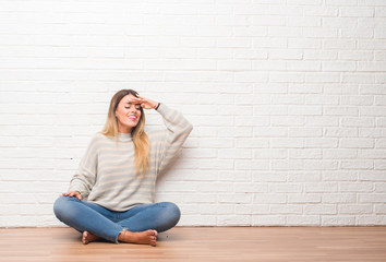 Young adult woman sitting on the floor over white brick wall at home very happy and smiling looking far away with hand over head. Searching concept.