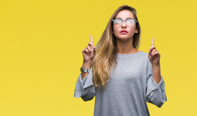 Young beautiful blonde woman wearing glasses over isolated background amazed and surprised looking up and pointing with fingers and raised arms.
