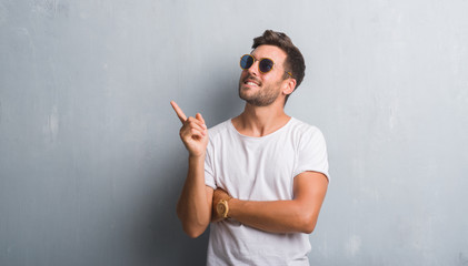 Handsome young man over grey grunge wall wearing sunglasses with a big smile on face, pointing with...