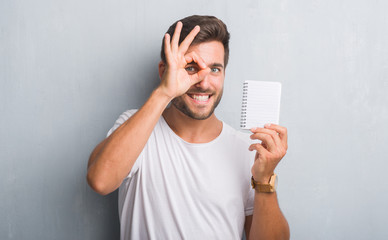 Handsome young man over grey grunge wall holding blank notebook with happy face smiling doing ok sign with hand on eye looking through fingers