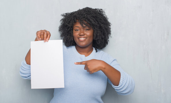 Young african american woman over grey grunge wall holding blank paper sheet very happy pointing with hand and finger