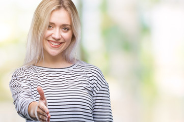 Young blonde woman over isolated background smiling friendly offering handshake as greeting and welcoming. Successful business.