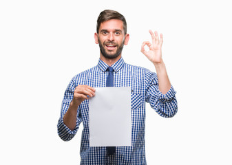 Young handsome man holding blank paper over isolated background doing ok sign with fingers,...