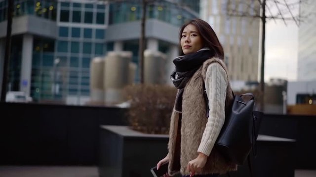 happy korean girl is strolling in city street in cold fall morning, looking around, touching hair