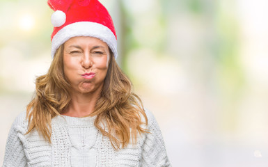 Middle age senior hispanic woman wearing christmas hat over isolated background puffing cheeks with funny face. Mouth inflated with air, crazy expression.