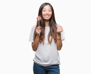 Young asian woman eating lollipop candy over isolated background screaming proud and celebrating victory and success very excited, cheering emotion