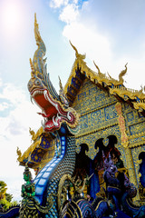 Thai Naga and angel statue is located in front of the temple of Thai architecture with sky and white clouds.