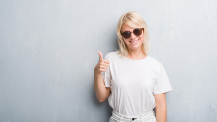 Adult caucasian woman over grunge grey wall wearing sunglasses doing happy thumbs up gesture with hand. Approving expression looking at the camera with showing success.