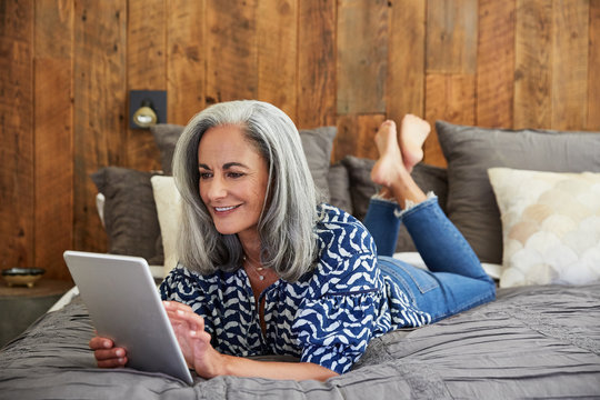 Mature woman with grey hair using a digital tablet on her bed at home smiling 