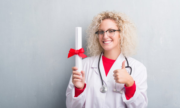 Young blonde doctor woman over grunge grey wall holding degree happy with big smile doing ok sign, thumb up with fingers, excellent sign