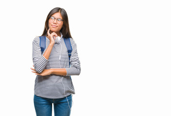 Young asian student woman wearing headphones and backpack over isolated background with hand on chin thinking about question, pensive expression. Smiling with thoughtful face. Doubt concept.