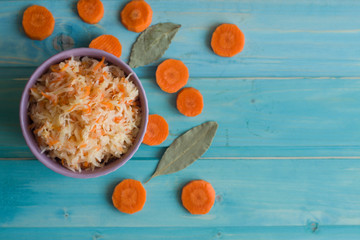 Photo of sauerkraut in a plate and round slices of carrots and bay leaf on a bright blue background.
