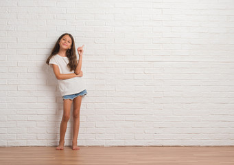 Young hispanic kid stading over white brick wall with a big smile on face, pointing with hand and...