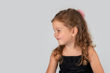 Waist up portrait of a little smiling girlwearing  beautiful black elegant dress.  She is siting against  white background in the studio and looking aside