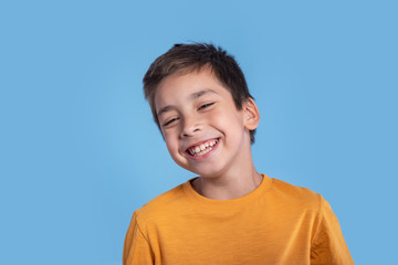 Close up emotional portrait of a  laughing boy wearing a yellow shirt with a mischievous or sly look on blue background in studio with copy space