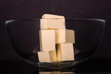 Pieces of butter lie in a glass bowl on a dark background in the studio.